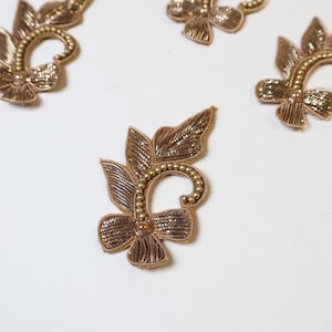 Peut inclure: Gros plan de plusieurs appliques florales de couleur bronze. Chaque applique présente un motif détaillé avec des feuilles, une fleur centrale et une tige incurvée. Les appliques sont ornées de petites perles dorées.