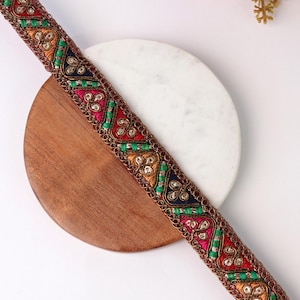 May include: A decorative fabric trim featuring a repeating pattern of triangular shapes in shades of red, orange, and dark blue. The design is accented with bright green thread details and metallic gold embroidery, finished with a scalloped border.