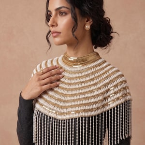 May include: A white and gold beaded cape with a high collar and fringe. The cape features horizontal rows of white beads and gold sequins, with long strands of white beads hanging from the bottom. The model is wearing a dark grey sweater.