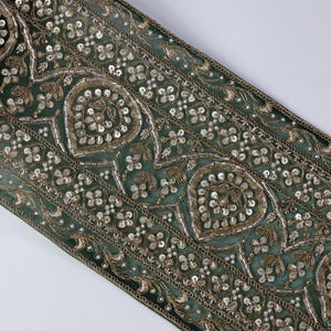 Può includere: Primo piano di una sezione di tessuto verde con ricami intricati. Il design presenta forme floreali e a goccia, impreziosite da paillettes argentate e filo dorato. I bordi sono rifiniti con un bordo dettagliato.