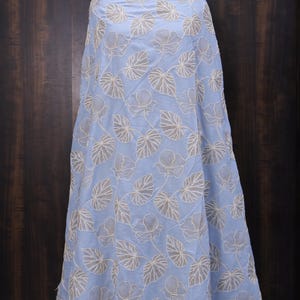 May include: Light blue fabric with an embroidered floral pattern. The design features white flowers and leaves. The fabric is draped over a form, showcasing the intricate details and texture of the embroidery.