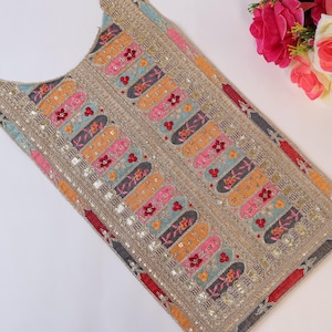 Puede incluir: Prenda bordada con base beige, con coloridos patrones verticales en rosa, naranja, azul y gris, acentuados con detalles florales rojos. Los bordes están adornados con adornos dorados. Un ramo de rosas rosas y blancas se encuentra en el fondo.