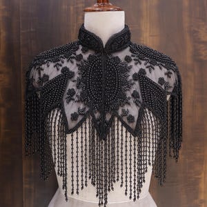 May include: A black beaded capelet with intricate floral and geometric patterns. The capelet features a high collar and long beaded fringe, displayed on a mannequin against a dark wood background. The capelet is made of black beads.