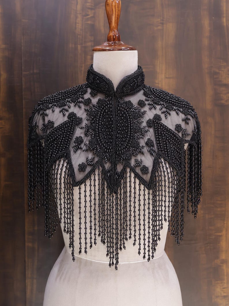 May include: A black beaded capelet with intricate floral and geometric patterns. The capelet features a high collar and long beaded fringe, displayed on a mannequin against a dark wood background. The capelet is made of black beads.