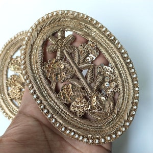 以下が含まれることがあります： 花柄の金色の楕円形の刺繍パッチ。デザインは中央の花と両側に2枚の葉があります。刺繍は白いビーズの縁で囲まれています。