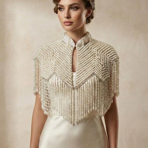 May include: Ivory-colored beaded capelet with a high collar and fringe detailing. The capelet is adorned with numerous small, round beads, creating a textured, elegant look. The design is suitable for bridal or formal wear.