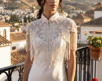 May include: A white wedding dress with a high neckline and a beaded, fringed overlay. The overlay features intricate floral embroidery and hangs over the shoulders. The dress is sleeveless and has a simple, elegant design.