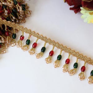 Può includere: Un bordo decorativo caratterizzato da una base in nastro intrecciato metallico dorato con frange pendenti. Le frange sono composte da perline ovali alternate rosse e verde scuro, abbinate a grappoli di piccoli cristalli color champagne traslucidi.