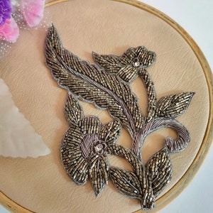 Peut inclure: Une applique florale en perles argentées avec un motif de feuille. L'applique est sur un fond beige.