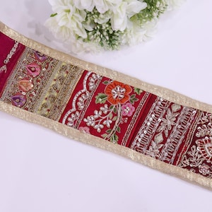 Può includere: Un bordo decorativo bordeaux con ricami e abbellimenti intricati. Il bordo presenta motivi floreali e geometrici in oro, rosa e argento, bordati da un bordo metallico dorato. Fiori bianchi sullo sfondo.