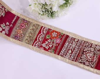 May include: A decorative trim featuring a rich burgundy base with intricate embroidery and embellishments. The trim showcases floral and geometric patterns in gold, pink, and silver, bordered by a gold metallic edge. White flowers are in the background.