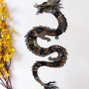 May include: A black, gold and silver sequined dragon applique. The dragon is curled in a serpentine shape with its head facing the left. The applique is perfect for adding a touch of whimsy and sparkle to clothing, bags or home decor.