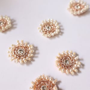 Könnte beinhalten: Eine Sammlung von acht kleinen, runden, dekorativen Verzierungen. Jede Verzierung besteht aus einer goldfarbenen Metallbasis mit einem Spiralmuster und ist von einem Ring aus weißen Perlen umgeben. Ein klarer Strassstein befindet sich in der Mitte jeder Verzierung.