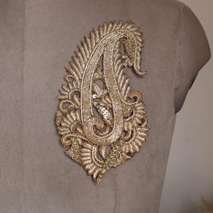 Pode incluir: Uma aplicação em forma de paisley de cor dourada com bordados intrincados de fio metálico e pequenas lantejoulas refletoras. O design apresenta um motivo de gota em espiral com costuras texturizadas detalhadas em toda a superfície.
