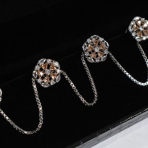 Puede incluir: Cuatro botones decorativos en tono plateado y oro rosa con diamantes de imitación transparentes y tono oro rosa, conectados por una cadena plateada.