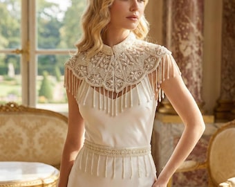 May include: Ivory-colored bridal gown with a high-collared, beaded capelet and a beaded belt. The capelet features intricate beadwork and dangling fringe. The dress has a simple, elegant design, perfect for a wedding or special occasion.