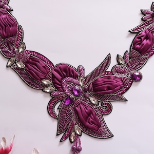 Pode incluir: Um enfeite de contas roxas e prateadas com um design floral. O enfeite é feito de tecido e contas e tem um tassel pendurado.