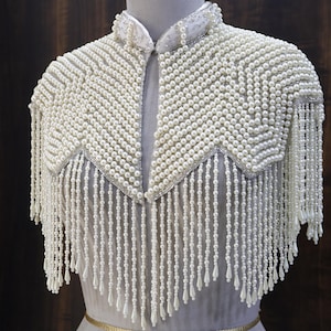 May include: A white beaded shoulder cape featuring a high collar and intricate rows of small round pearls arranged in a chevron pattern. The garment is adorned with long, dangling pearl fringe strands that end in teardrop-shaped beads.