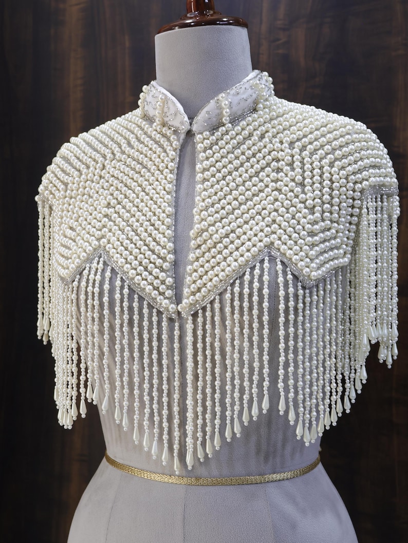 May include: A white beaded shoulder cape featuring a high collar and intricate rows of small round pearls arranged in a chevron pattern. The garment is adorned with long, dangling pearl fringe strands that end in teardrop-shaped beads.