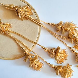 Pode incluir: Borlas decorativas douradas com intrincados trabalhos de contas e detalhes florais. As borlas são suspensas de uma corrente dourada e dispostas sobre uma superfície creme, criando um design elegante e ornamentado.