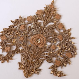 Puede incluir: Una aplicación bordada en tonos dorados que presenta un motivo floral central rodeado de intrincados patrones de hojas. El diseño está adornado con hilos metálicos, texturas de tela beige en relieve y pequeñas lentejuelas doradas.