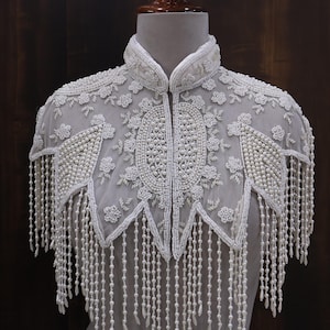 May include: A white beaded cape with a high collar and intricate floral and geometric patterns. The cape features long beaded fringe and is displayed on a mannequin. The design is elegant and detailed.