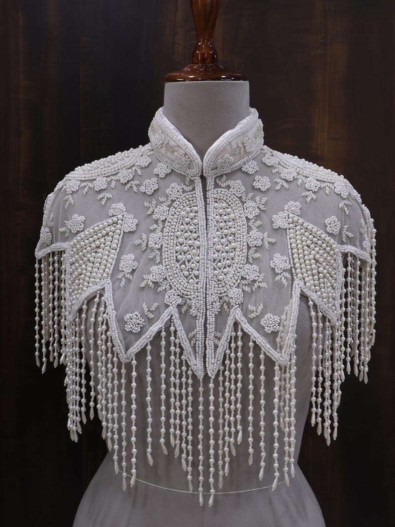 May include: A white beaded cape with a high collar and intricate floral and geometric patterns. The cape features long beaded fringe and is displayed on a mannequin. The design is elegant and detailed.