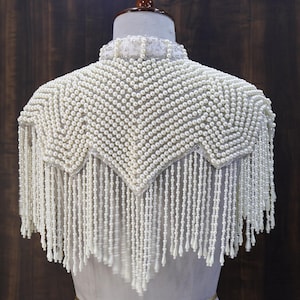 May include: A white pearl-beaded shoulder cape featuring a high collar and a chevron-patterned yoke. The garment is adorned with long, dangling strands of white pearls that form a fringe, creating a layered texture across the shoulders.