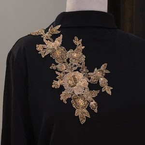 Può includere: Camicia nera con colletto con ricamo floreale dorato sulla spalla. Il ricamo presenta dettagli di perline e paillettes, creando un aspetto lussuoso. La camicia è esposta su un manichino.