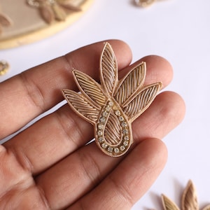 May include: A gold-colored embroidered applique with a teardrop shape and a center of clear rhinestones. The applique has a floral design with multiple layers of fabric.