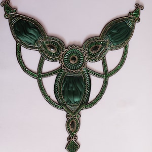 Puede incluir: Una decoración de cuentas verdes y doradas con un diseño central que presenta una flor de tela verde. La decoración tiene un borla colgante con cuentas verdes.