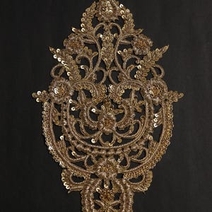 Può includere: Un'applicazione ricamata color oro con intricati lavori in filo metallico e paillettes scintillanti disposte in un motivo floreale e a volute simmetrico e ornato. L'elemento decorativo presenta una parte superiore e inferiore appuntita.