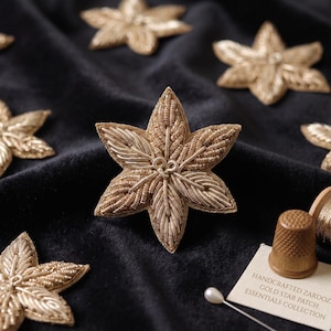 May include: Multiple six-pointed star patches made of gold metallic thread and beads. A small wooden thimble, a spool of gold thread, and a white card with the text "HANDCRAFTED ZARDOZI GOLD STAR PATCH ESSENTIALS COLLECTION" are visible.