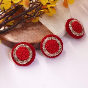 May include: Three red, round earrings with a textured center and a silver-colored, rope-like border. The earrings are displayed on a white surface with a wooden and marble accent, and yellow flowers.