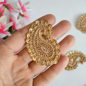 May include: Gold-colored paisley applique with intricate embroidery and sequin detailing. The teardrop-shaped design features a central beaded section and a decorative border. Additional gold appliques are visible in the background, suggesting a craft or embellishment theme.