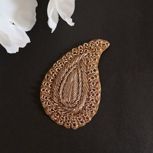 May include: A gold-colored paisley-shaped embellishment with a leaf design in the center. The embellishment is made of intricate metalwork and is perfect for adding a touch of elegance to clothing or crafts.