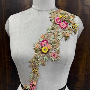 Op de afbeelding: Een bloemenapplicatie met roze, gele en groene bloemen en gouden pailletten. De applicatie is op een wit mannequin.