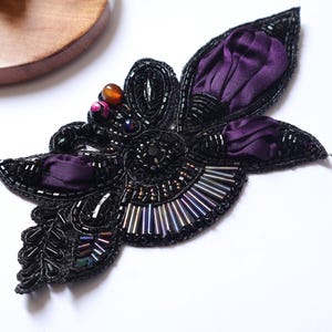 May include: A black beaded floral embellishment with purple fabric accents. The flower-shaped design features intricate beadwork, including small black beads and larger, colorful beads. The petals are made of a deep purple fabric.