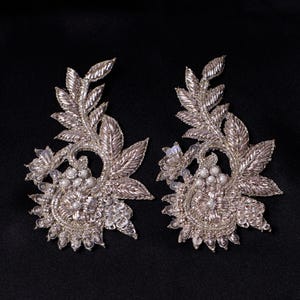 Puede incluir: Dos apliques florales bordados en color plata. Cada aplique presenta un diseño detallado con hojas, detalles de cuentas y un grupo central de pequeñas perlas redondas. Los apliques están sobre un fondo oscuro.