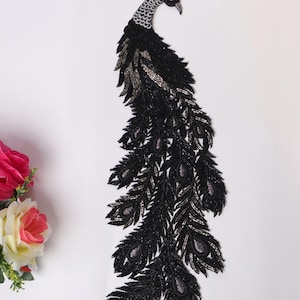 May include: A black and silver beaded peacock applique. The peacock has a detailed design with a fan of feathers. The head is adorned with a small red jewel. The applique is suitable for embellishing clothing or accessories.