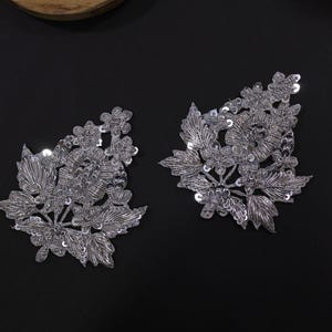Peut inclure: Deux écussons floraux argentés avec des détails complexes. Chaque écusson présente un motif de feuilles, de fleurs et de paillettes. Les écussons sont placés sur une surface noire, créant un contraste saisissant. Idéal pour embellir des vêtements ou des créations.