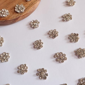 May include: Close-up of several silver-colored flower-shaped embellishments with clear crystal-like accents. Some are scattered on a white surface, while others rest on a wooden tray. These decorative elements are likely for crafts or embellishing clothing.