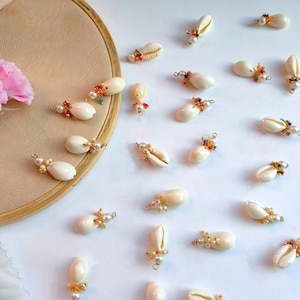 Könnte beinhalten: Eine Sammlung von weißen Muschelanhängern mit goldenen Akzenten und bunten Perlen. Die Anhänger sind auf einer weißen Oberfläche angeordnet.