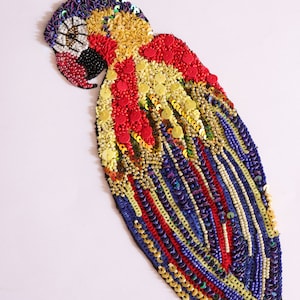 Può includere: Un'applicazione di perline a forma di pappagallo colorata con paillettes rosse, gialle, blu e dorate. L'applicazione è realizzata con perline e paillettes cucite su una base di tessuto.