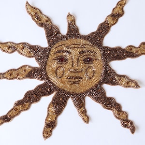 May include: A sun-shaped appliqué patch crafted with intricate beadwork in shades of metallic gold and deep bronze. The central face features a serene expression with defined eyes and cheeks, surrounded by twelve wavy, pointed rays extending outwards.