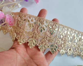 May include: Gold-colored embroidered lace trim with a floral and leaf design. The trim features a scalloped edge and is approximately 2 inches wide. The lace is held in a hand, with pink and purple flowers in the background.