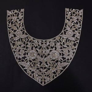 Op de afbeelding: Zilveren geborduurde bloemenkraag met ingewikkelde details. De kraag heeft een U-vormig ontwerp met een dicht patroon van bloemen, bladeren en pailletten. De borduursels zijn gemaakt met zilveren draad, wat een delicate en elegante uitstraling creëert. Op een zwarte achtergrond.