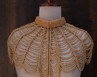 Puede incluir: Collar de cuentas doradas exhibido en un maniquí. El collar presenta un cuello alto y redondeado y un diseño tipo capa, con hilos de cuentas que caen en cascada. Las cuentas son de color dorado uniforme, creando un efecto brillante.