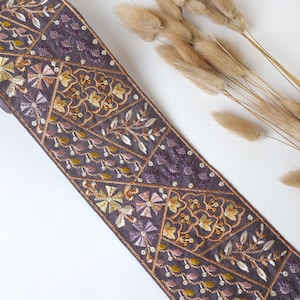 以下が含まれることがあります： 紫色の生地のトリムに、金色とピンク色の刺繍が施されています。トリムは、花柄の幾何学模様になっています。
