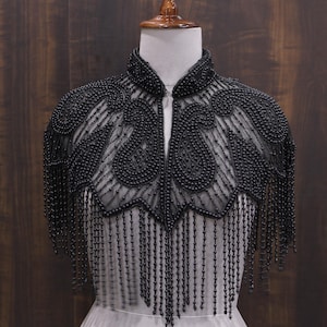 May include: A black beaded capelet with a high collar and intricate paisley-like designs. The capelet features long beaded fringe that hangs down from the shoulders. The garment is displayed on a mannequin against a dark wood background.